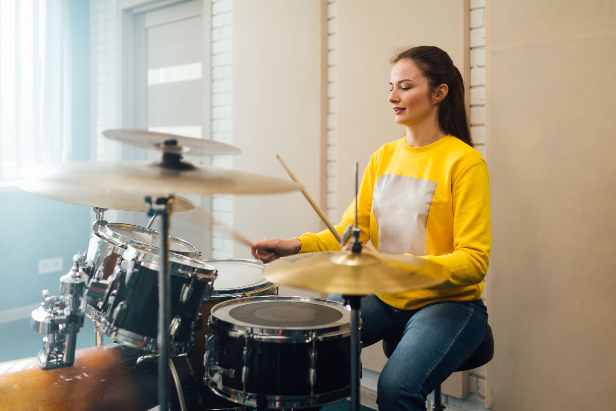 A woman wearing a yellow sweater plays a drum kit, smiling gently while holding drumsticks over the cymbals and snare.