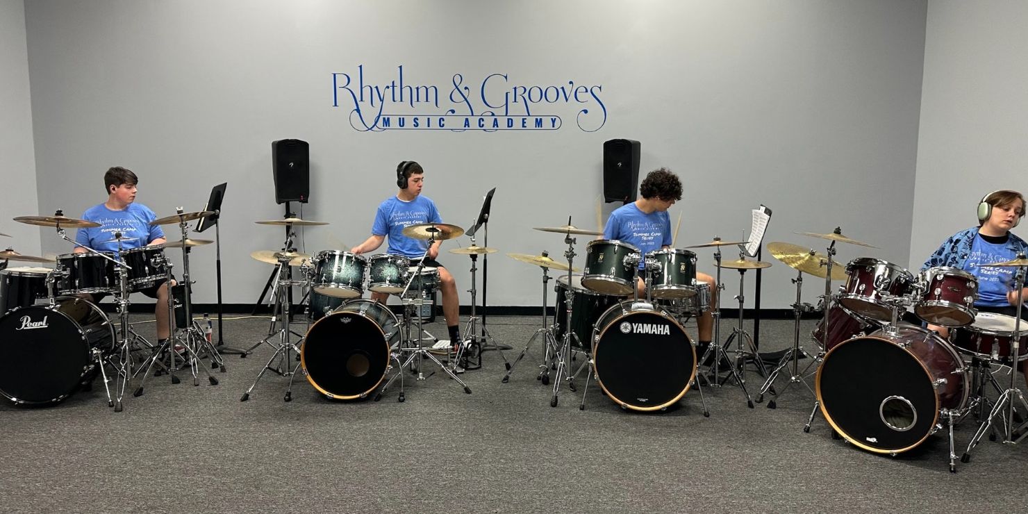 Four students in matching blue camp t-shirts play drum kits in a row at Rhythm & Grooves Music Academy, with the academy's logo on the wall behind them.