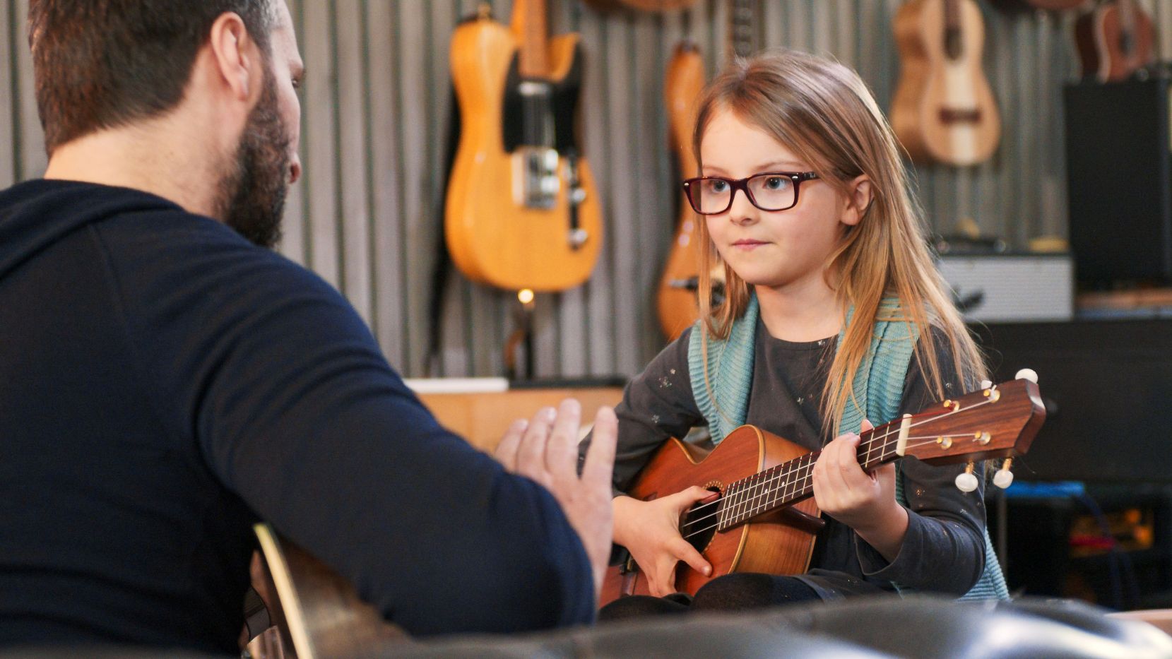 young girl wearing glasses holds a ukulele and listens attentively to an instructor, with several guitars and ukuleles displayed on the wall behind her.