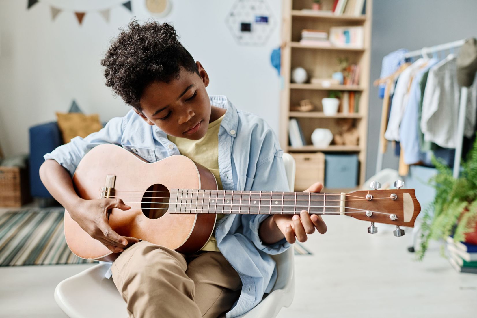 A young boy with curly hair, wearing a light blue button-up shirt, plays a ukulele while sitting in a bright bedroom with a bookshelf in the background.