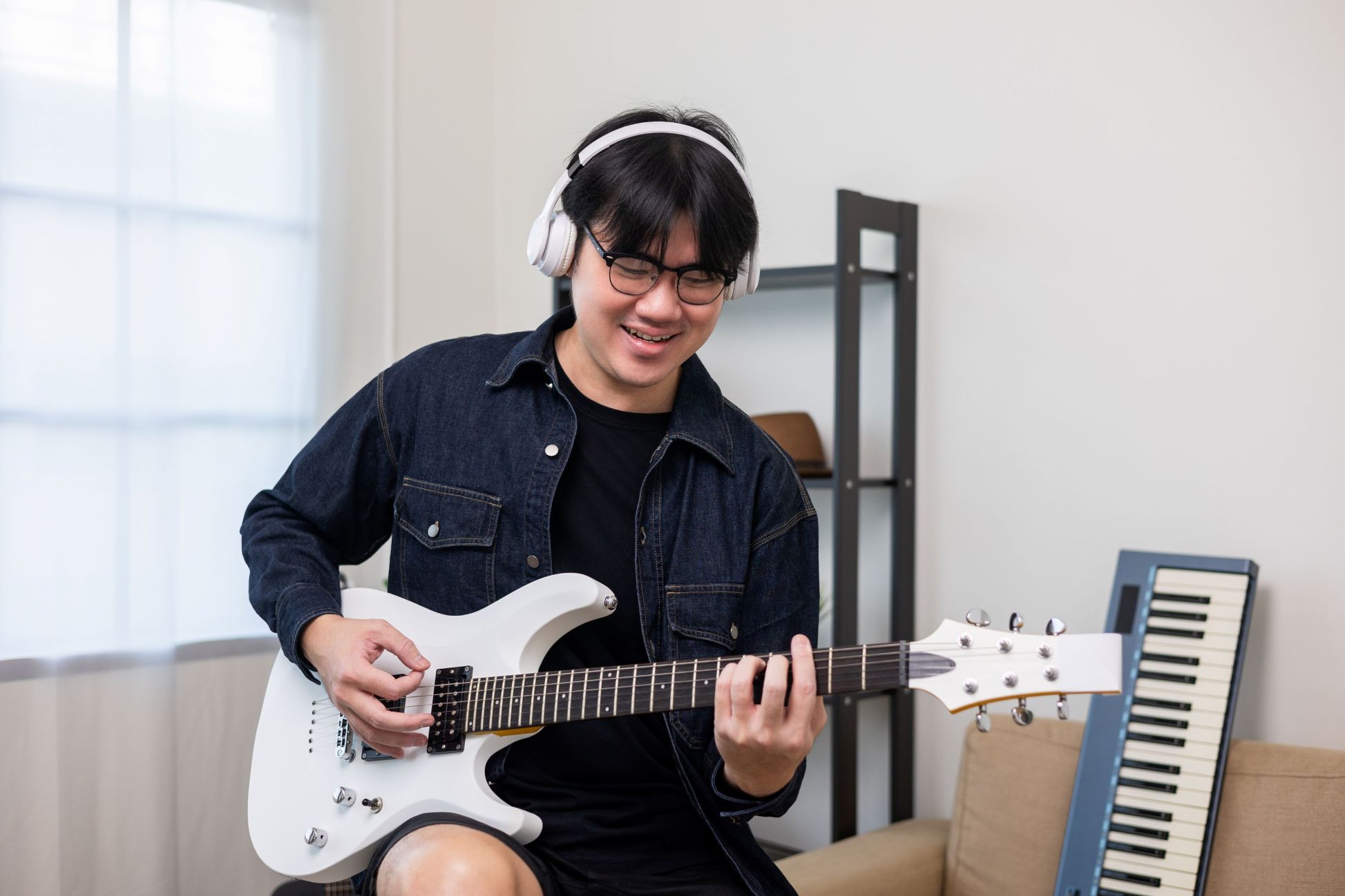 A man wearing glasses, headphones, and a denim jacket smiles while playing a white electric guitar in a bright indoor setting.