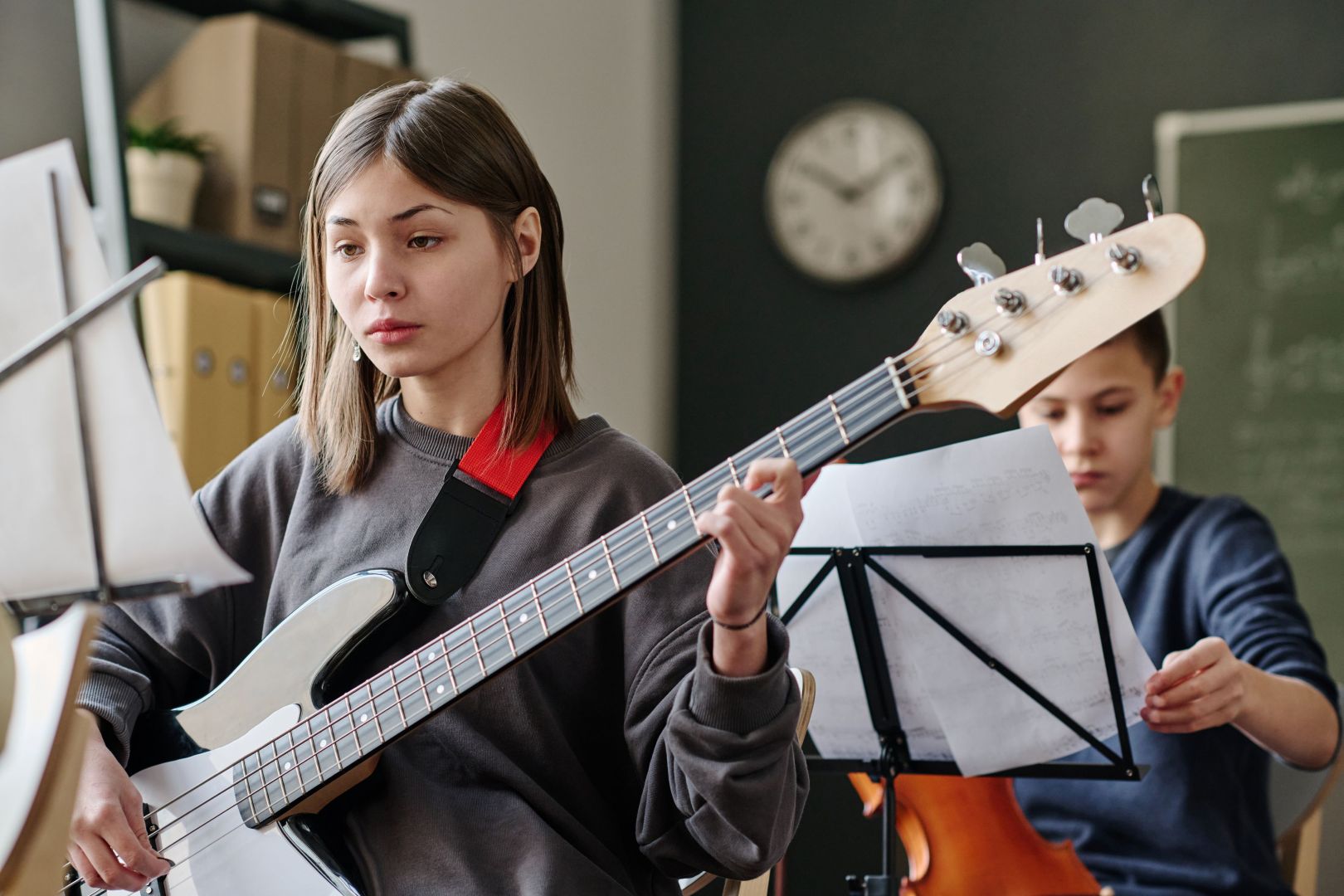 A young woman plays an electric bass guitar while reading sheet music, with a boy holding music sheets in the background and a clock on the wall.