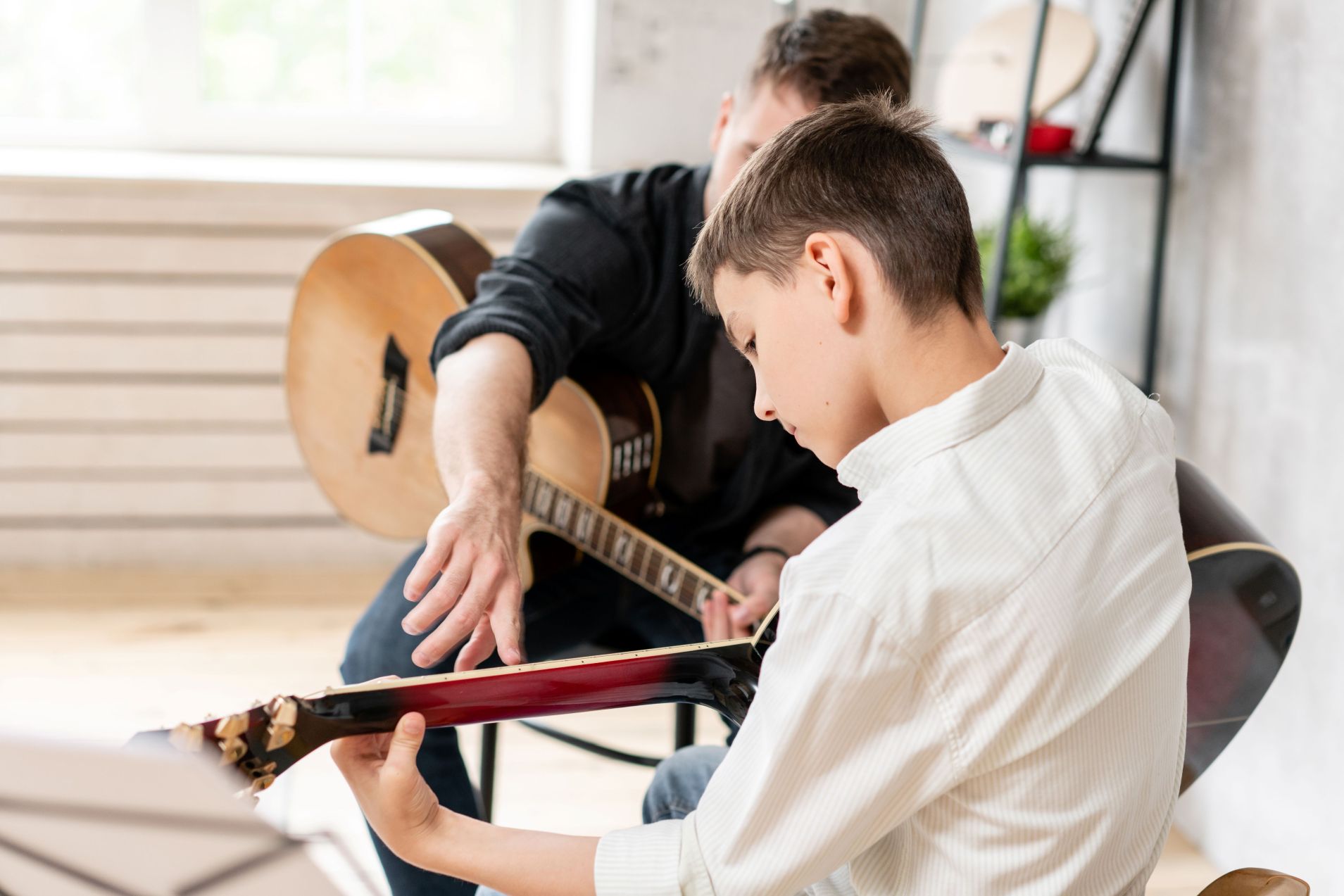 A young boy holds a red electric guitar while an instructor with an acoustic guitar gestures, demonstrating technique during a lesson.