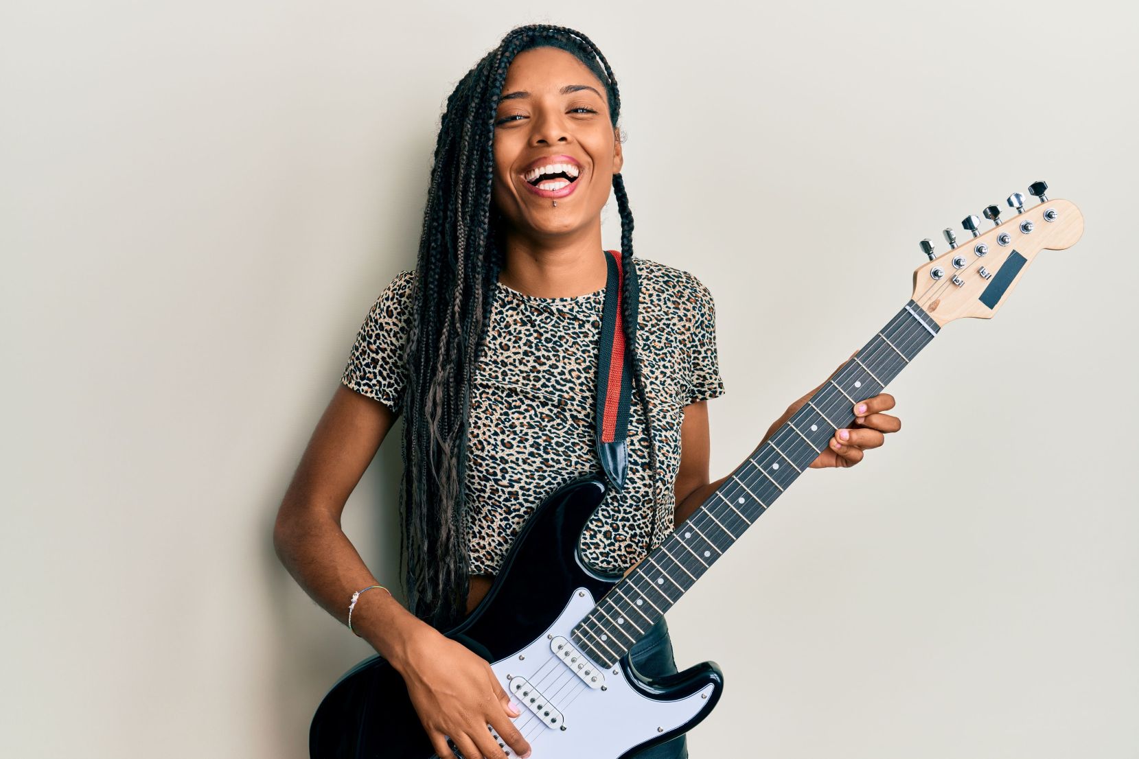 A woman with long braided hair, wearing a leopard-print top, laughs joyfully while holding a black electric guitar against a plain beige background.