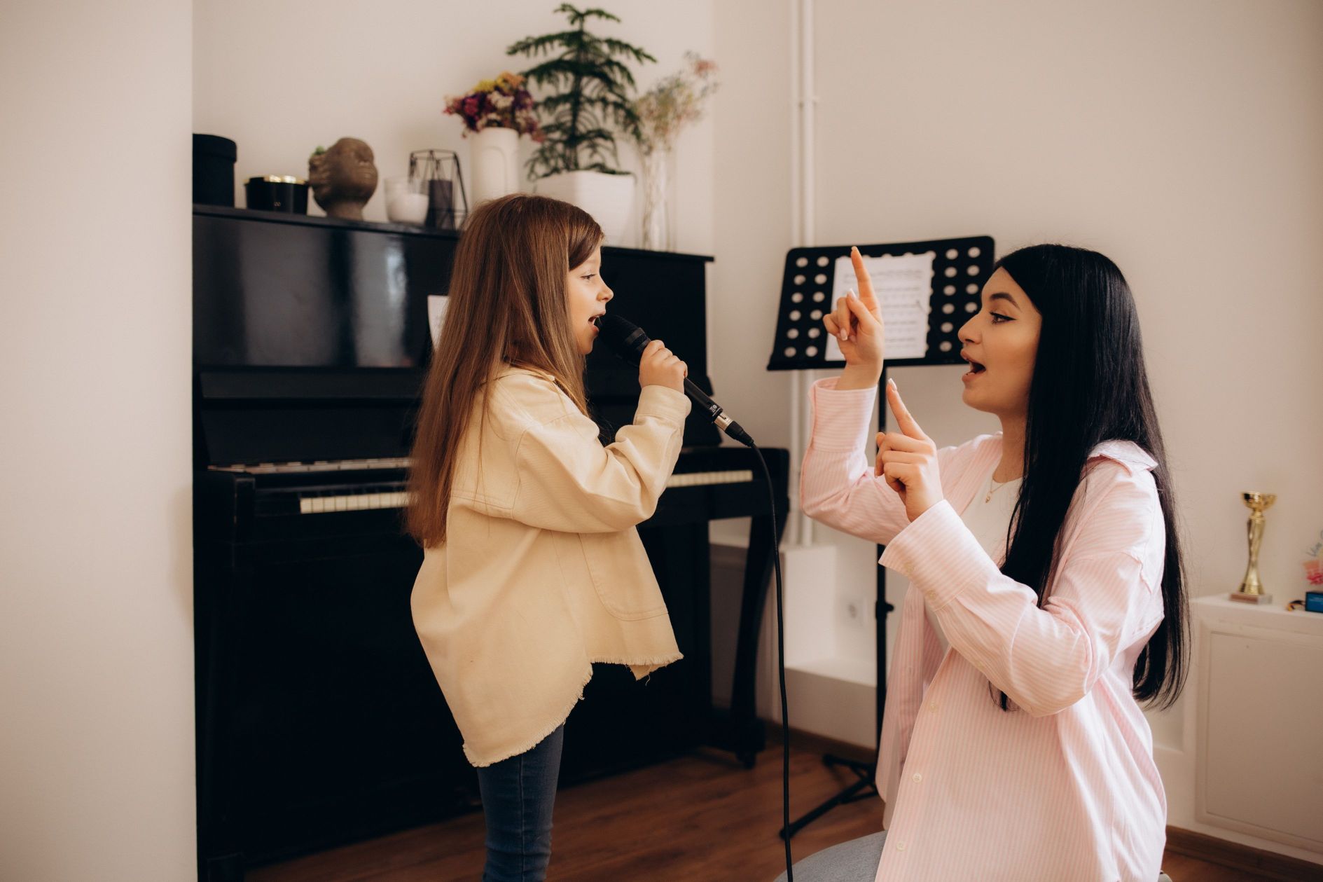 A vocal instructor gestures while guiding a young girl singing into a microphone, standing near a piano and sheet music stand.