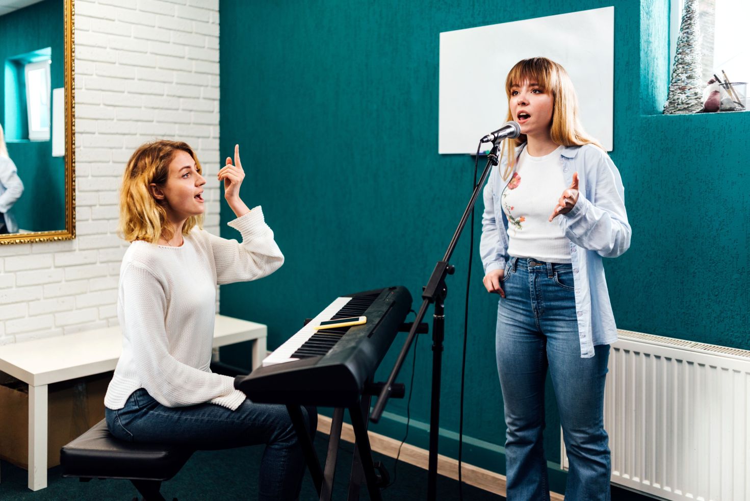 A vocal coach at a keyboard gestures while instructing a young woman who sings into a standing microphone, in a room with teal walls and a whiteboard.