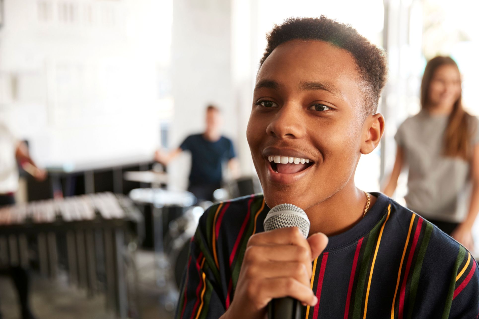 A young man wearing a striped shirt smiles while singing into a microphone, with a drummer visible playing in the blurred background.