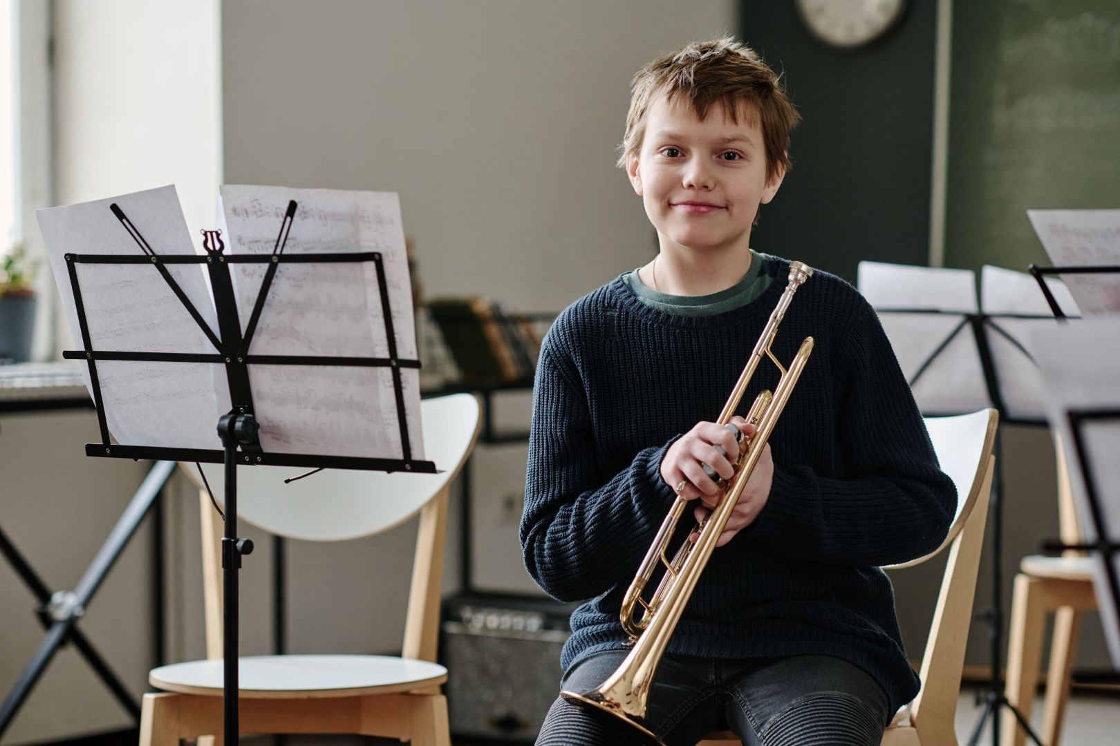 A boy wearing a navy sweater sits and smiles at the camera while holding a trumpet, with music stands and sheet music around him in a classroom.