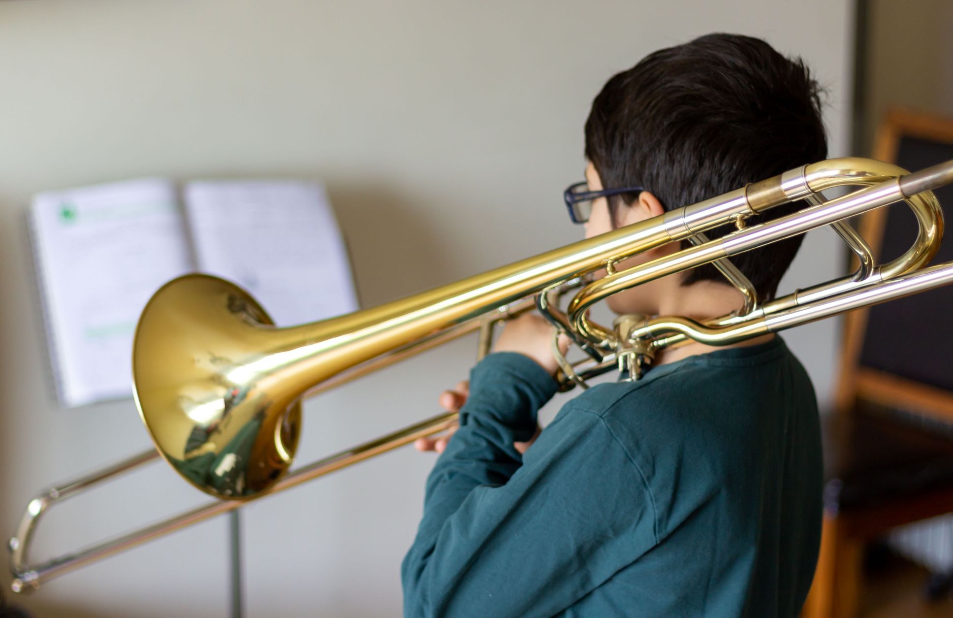 A boy wearing glasses plays the trombone, seen from behind, with sheet music on a stand in the background.