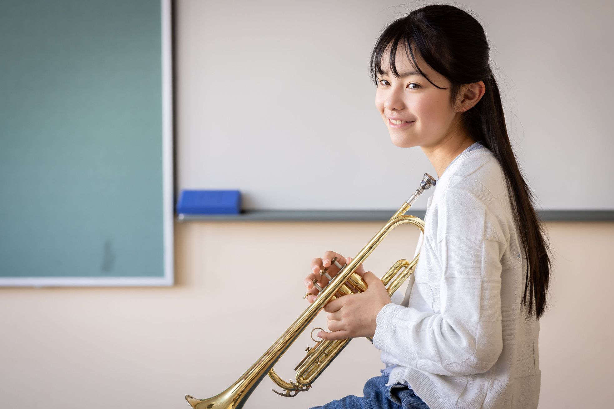 A girl with long dark hair smiles at the camera while holding a trumpet, sitting in a classroom with a chalkboard in the background.