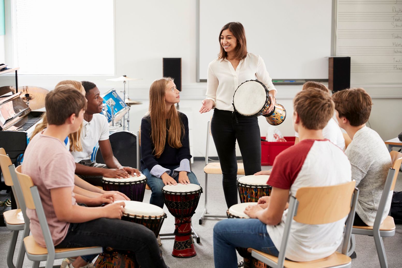 A teacher holding a djembe drum leads a group class, with students seated in a circle each playing their own djembe drums.