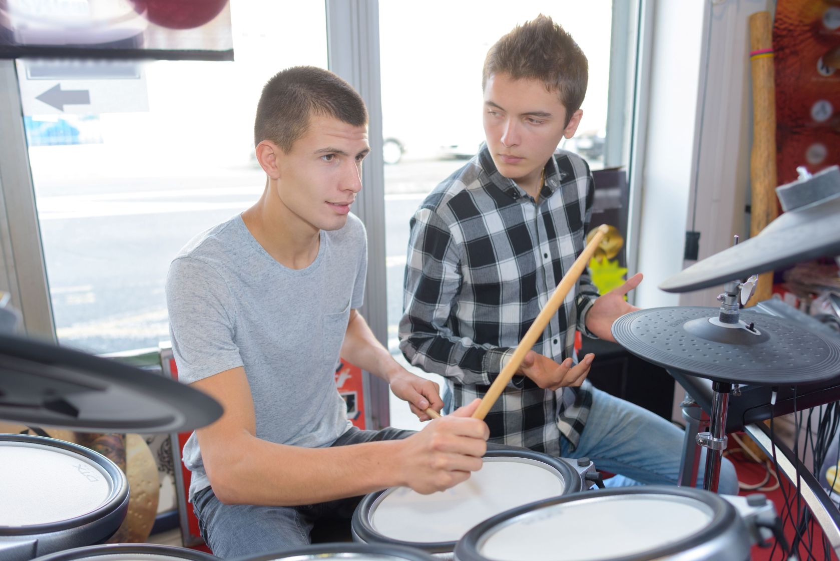 Two young men sit at an electronic drum kit in a music shop, one holding a drumstick while the other gestures as they talk.