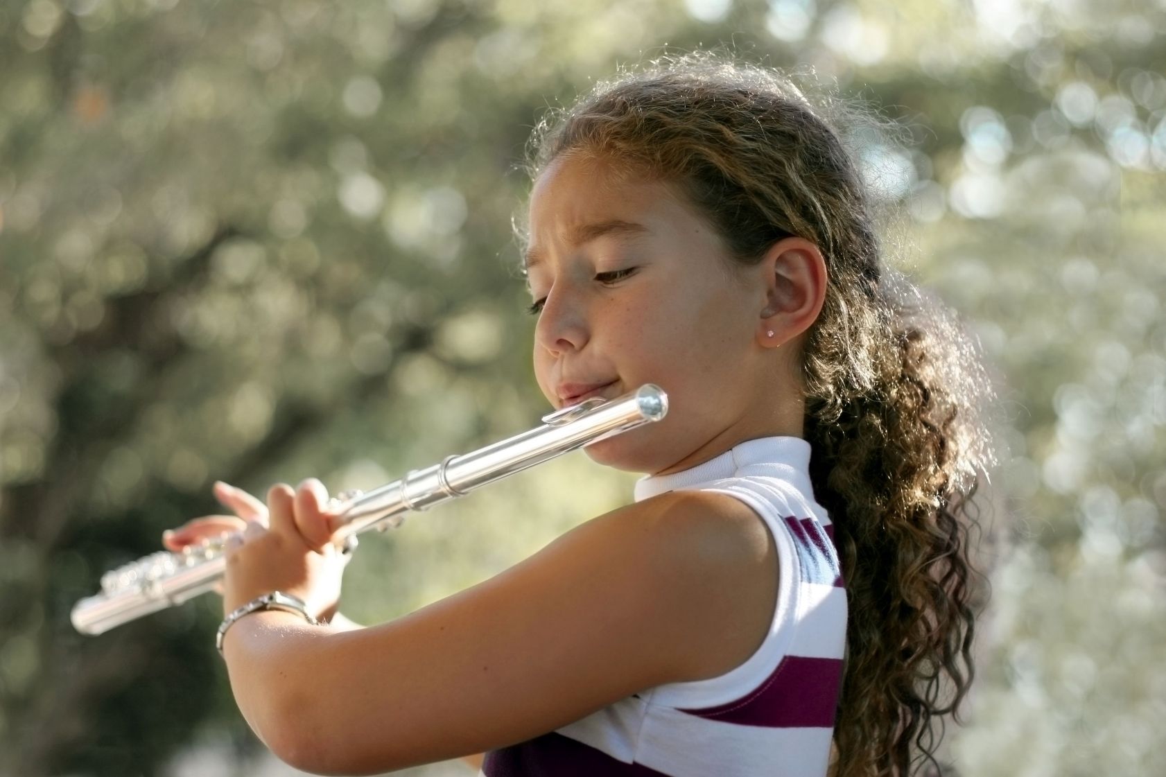A young girl with curly hair plays the flute outdoors, eyes closed in concentration, with sunlight filtering through trees in the background.