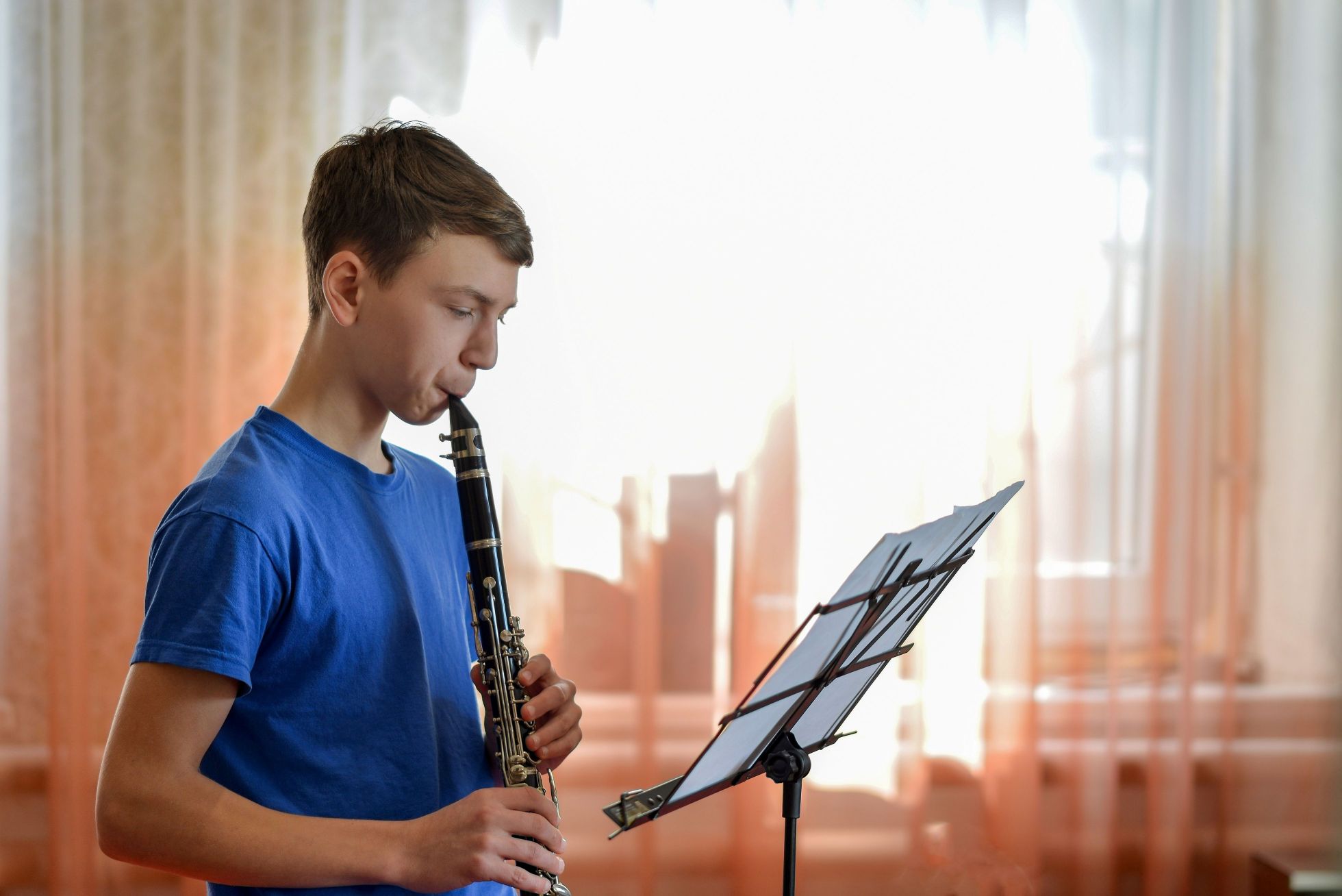 A teenage boy wearing a blue t-shirt plays the clarinet, reading sheet music on a stand near sunlit sheer curtains.