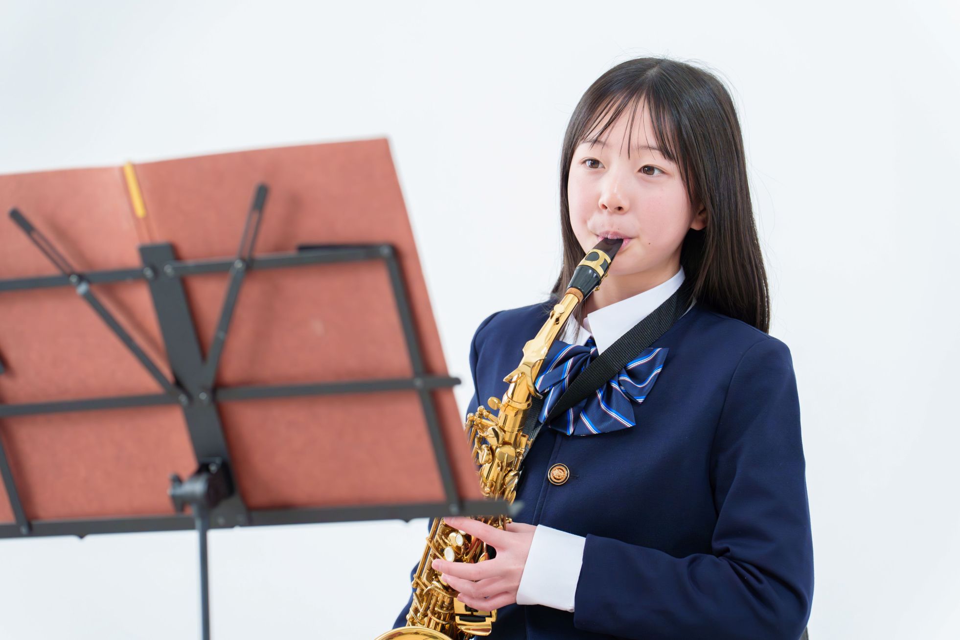A girl in a school uniform plays a saxophone beside a music stand, against a plain white background.