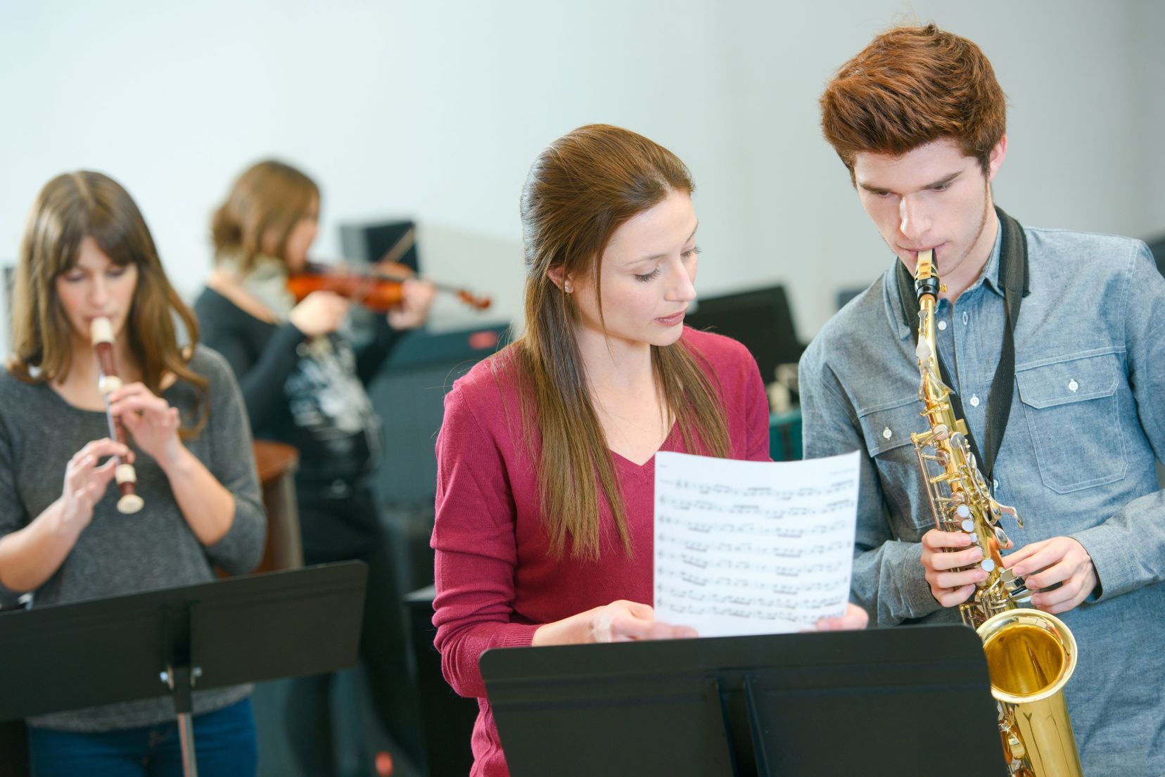 A man plays saxophone while a woman holds up sheet music beside him, with two more musicians playing recorder and violin in the background.