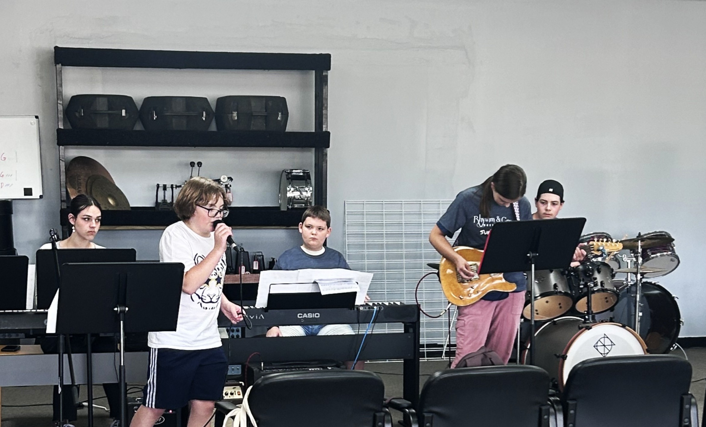 A boy sings into a microphone beside a keyboard player, while another student plays electric guitar near a drum kit in a band rehearsal room.