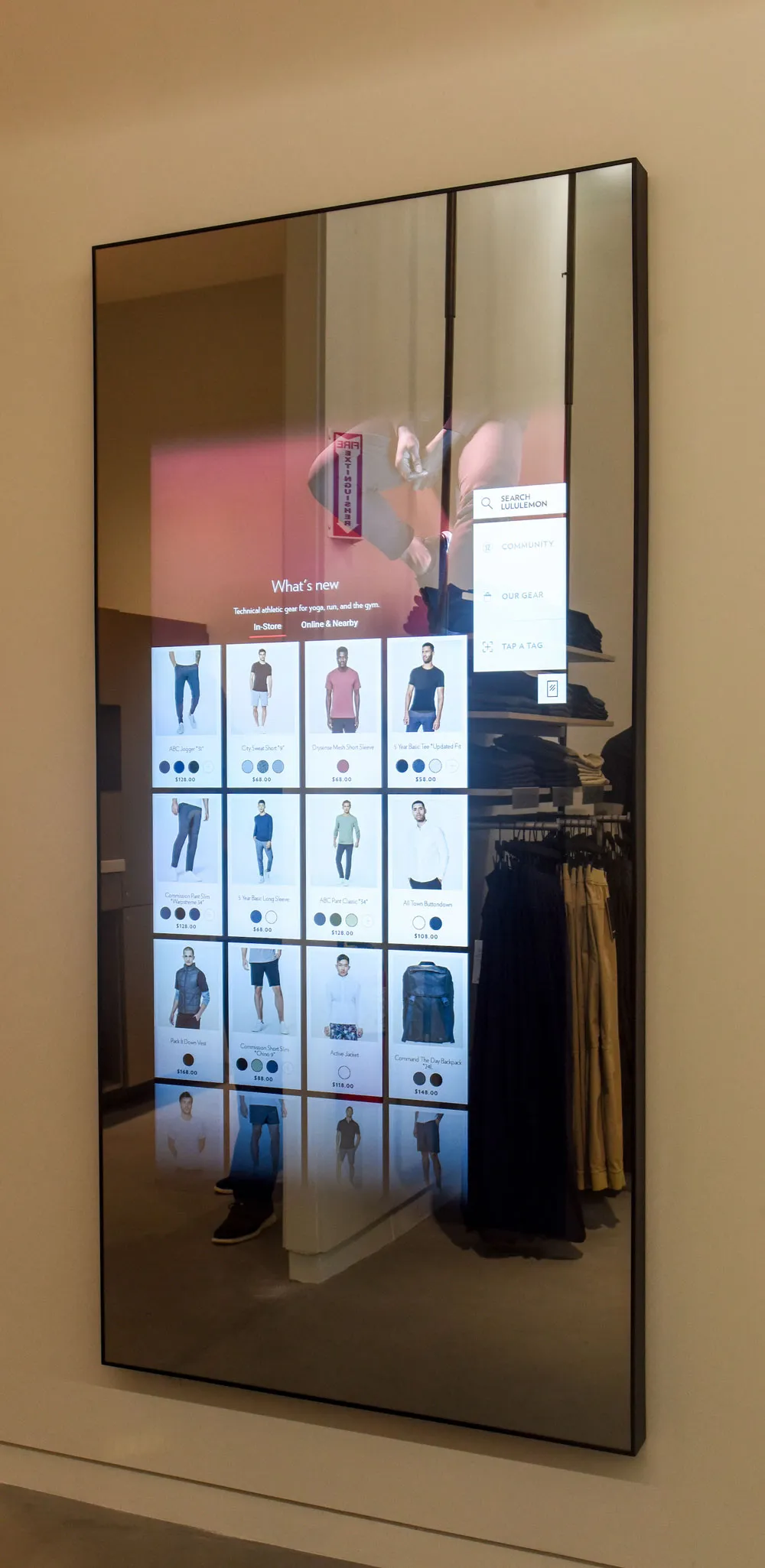 A full-length mirror displaying an interactive digital interface with clothing options organized in a grid. The background features a clothing display rack in a retail store.