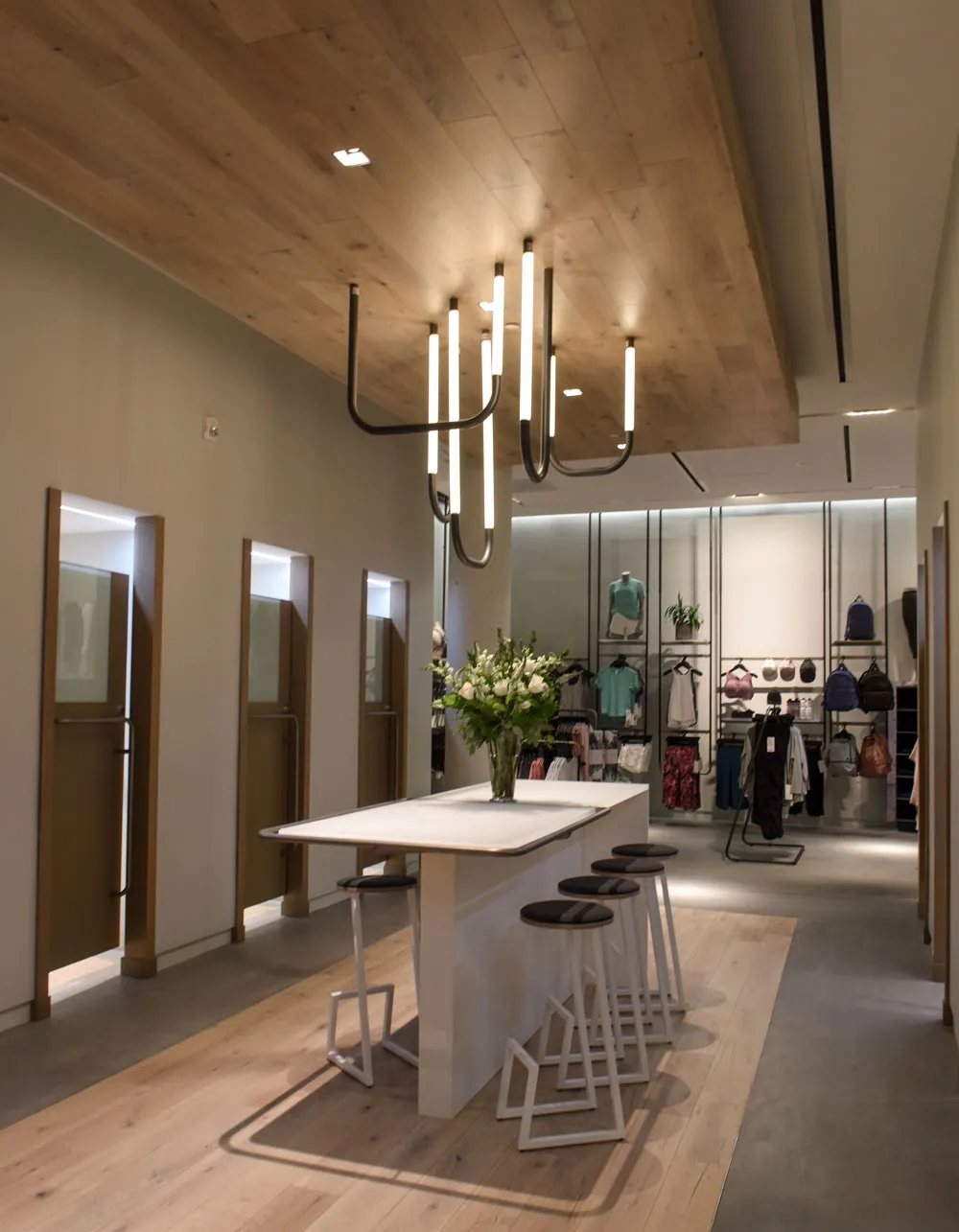 A modern retail space with wooden flooring, featuring a stylish chandelier and a central table with stools. Clothing and accessories are displayed on racks in the background, and floral arrangements are placed on the table.