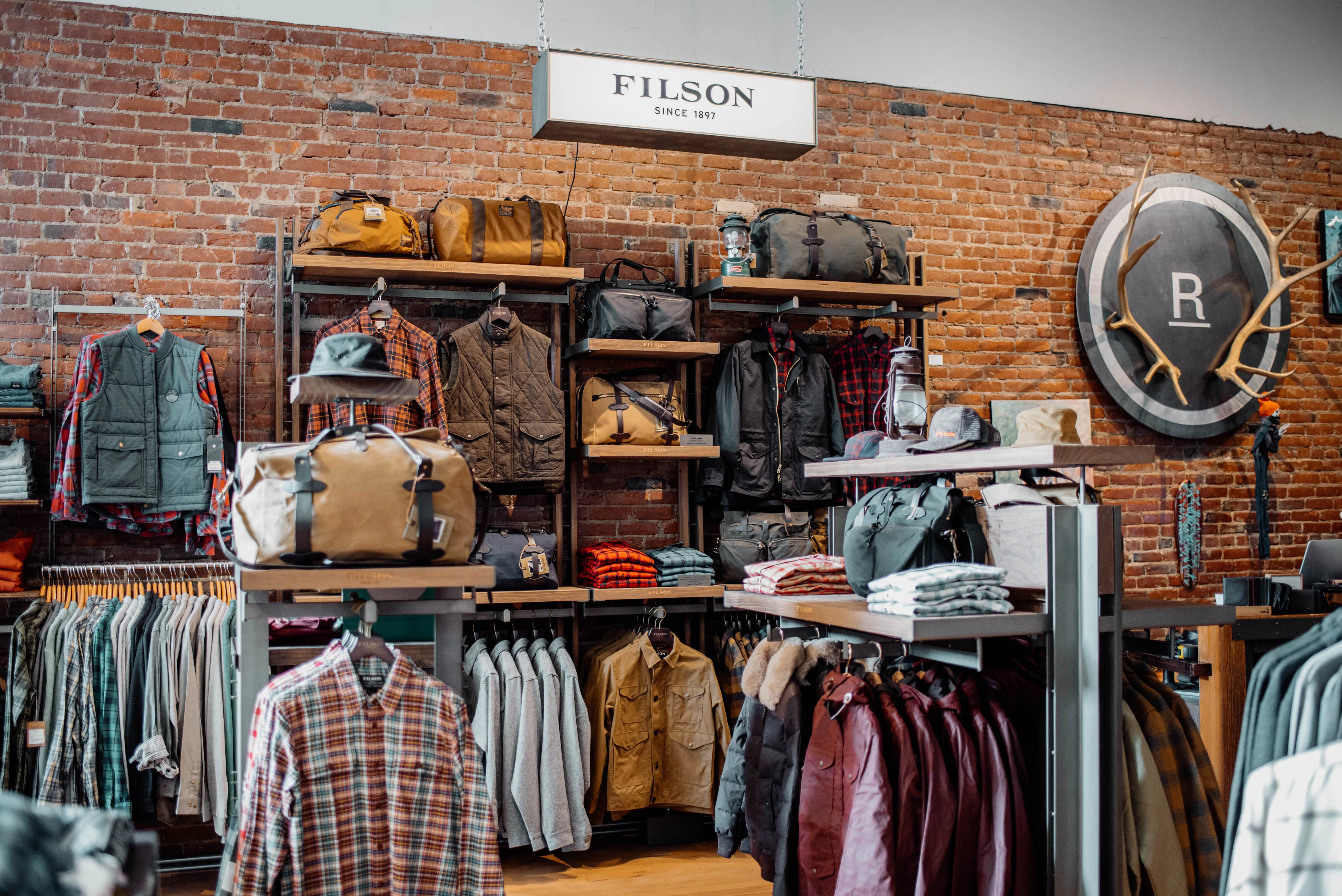 FILSON retail store display featuring exposed brick wall with organized merchandise including plaid shirts, leather bags, jackets, and outdoor apparel on metal shelving and clothing racks with branded signage