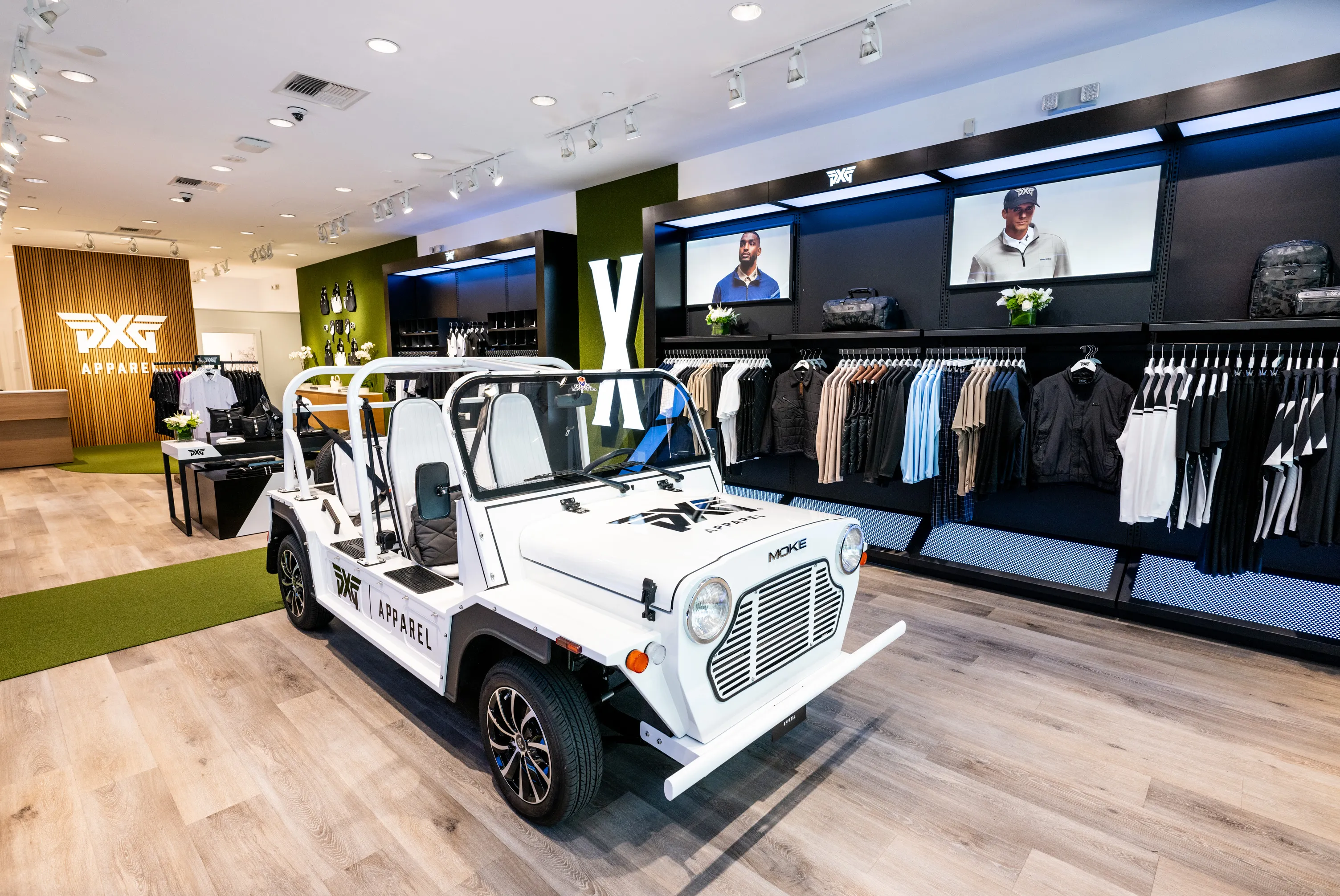 An indoor retail store featuring a collection of apparel on racks and displayed on screens. A small white vintage-style car with 'PXG Apparel' branding is positioned in the center of the store. The interior has light wood flooring and modern lighting.