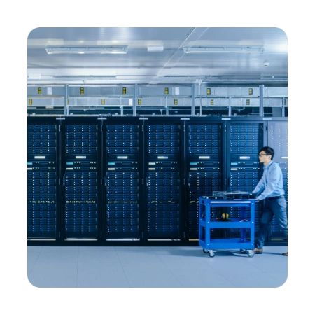 An IT technician pushing a blue equipment trolley through a modern, high density data centre aisle lined with tall server racks displaying blue LED indicator lights.