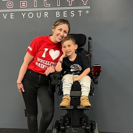 Smiling woman in red t-shirt standing next to young boy sitting in a motorized wheelchair against a gray wall.