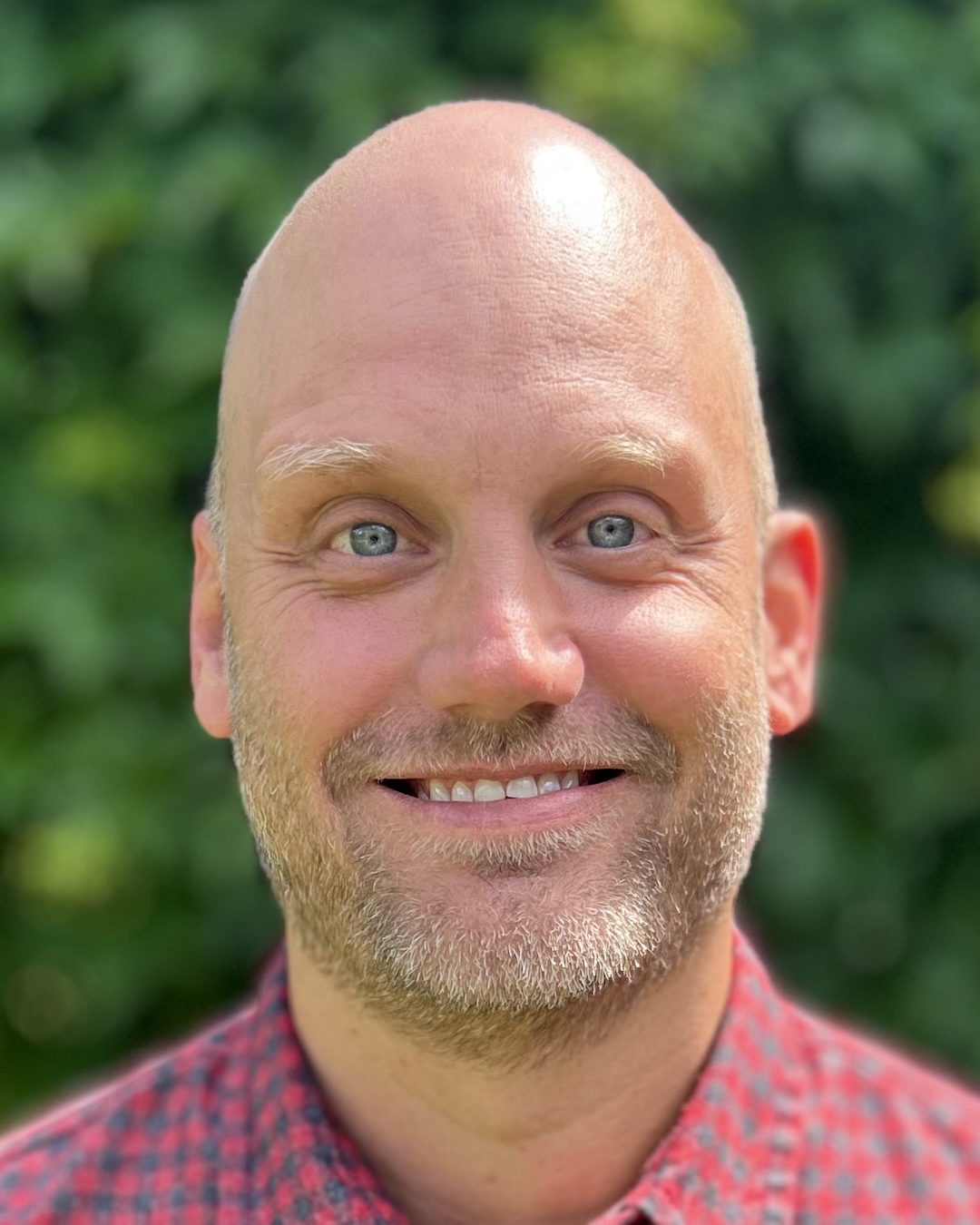 Smiling bald man with blue eyes and light beard wearing a red checkered shirt against a green leafy background.