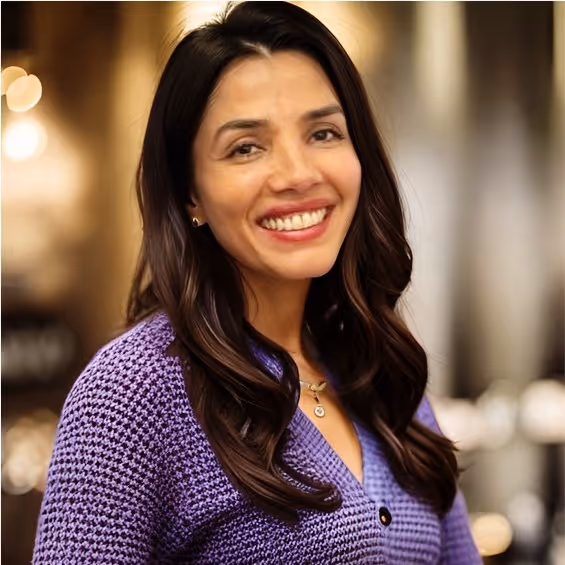 Smiling woman with long dark hair wearing a purple textured sweater and a necklace, in a softly lit indoor setting.