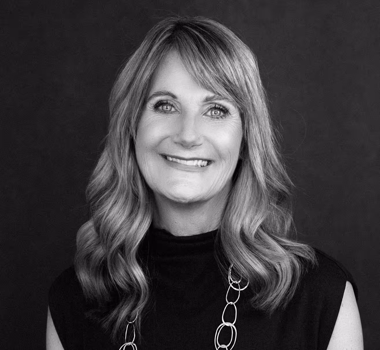 Smiling woman with wavy shoulder-length hair wearing a black sleeveless top and a chain-link necklace, against a dark background.