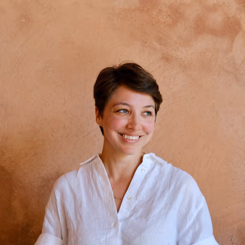 Smiling woman with short brown hair wearing a white shirt against a textured tan background.