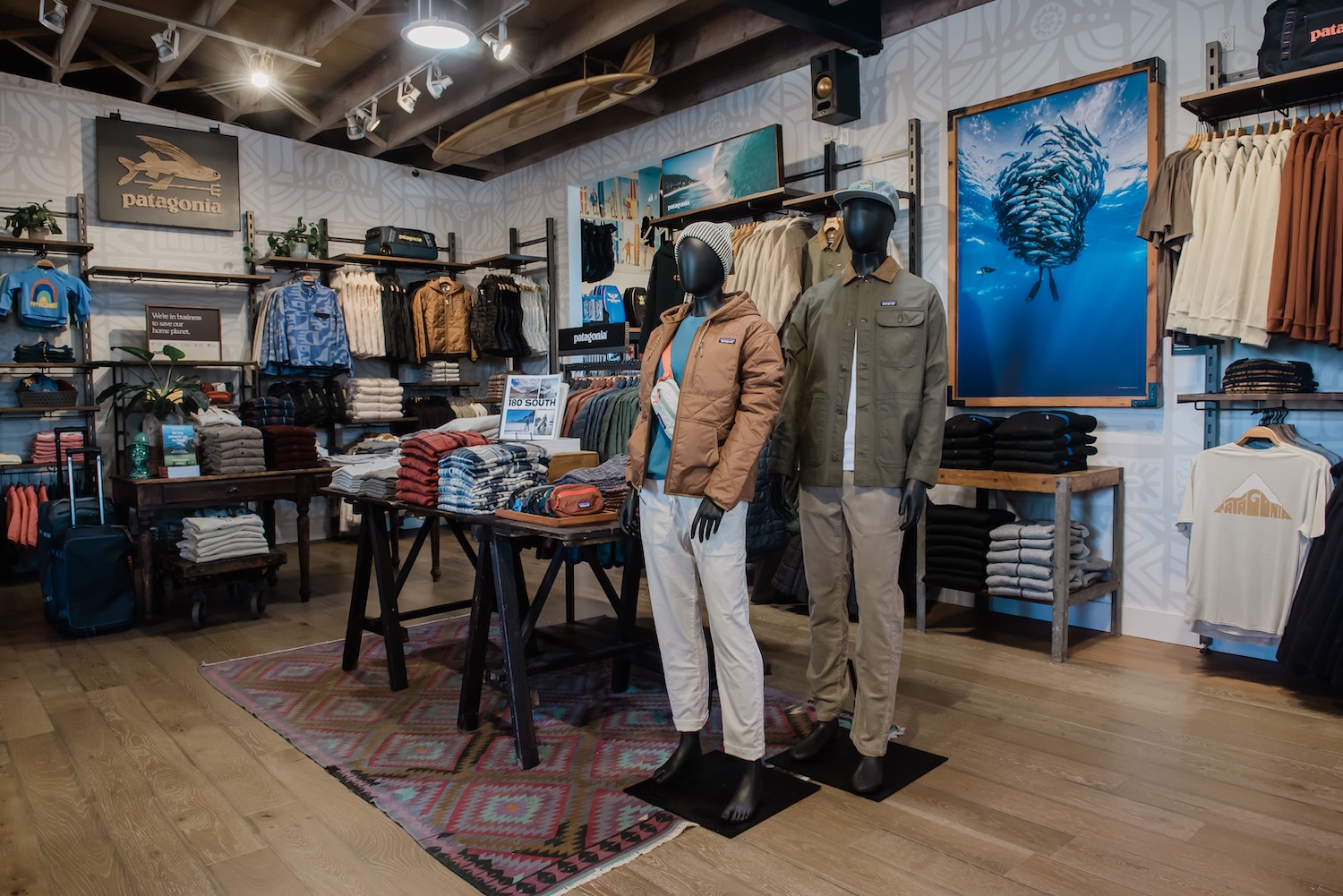 Interior of a Patagonia retail store with warm wood floors, open metal-and-wood wall fixtures, folded apparel on rustic tables, and two mannequins in outdoor wear.