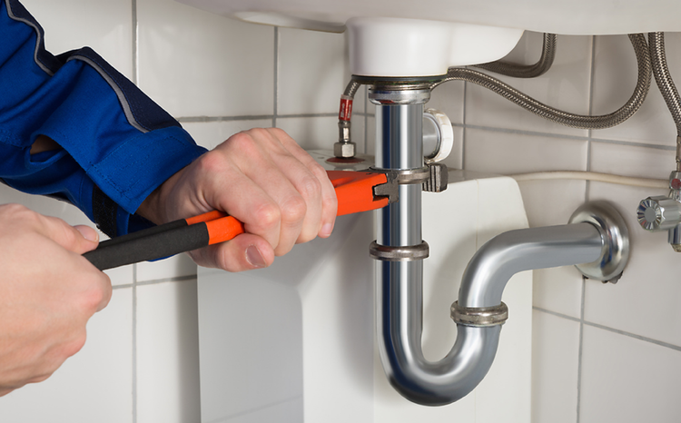 Hands using an orange pipe wrench to tighten a chrome drain pipe under a sink.
