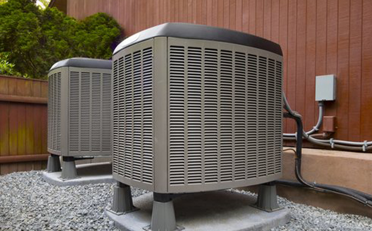 Two outdoor air conditioning units installed on concrete pads beside a wooden building wall.