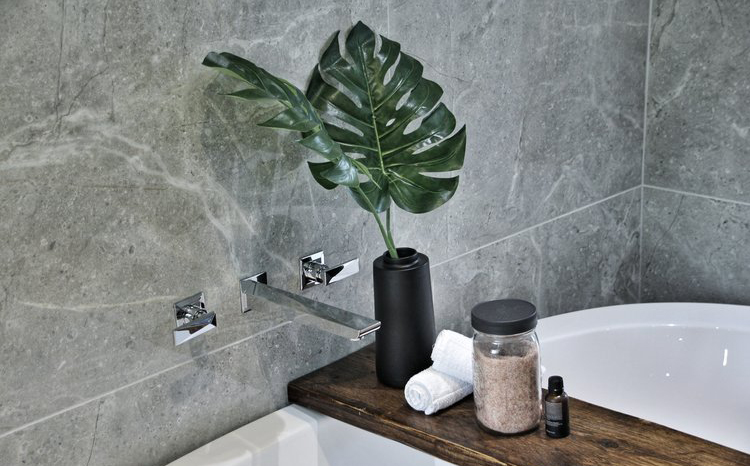 Modern bathroom corner with gray marble walls, a white bathtub, a wooden shelf holding a black vase with large green leaves, rolled white towels, a jar of bath salts, and a small dark bottle.