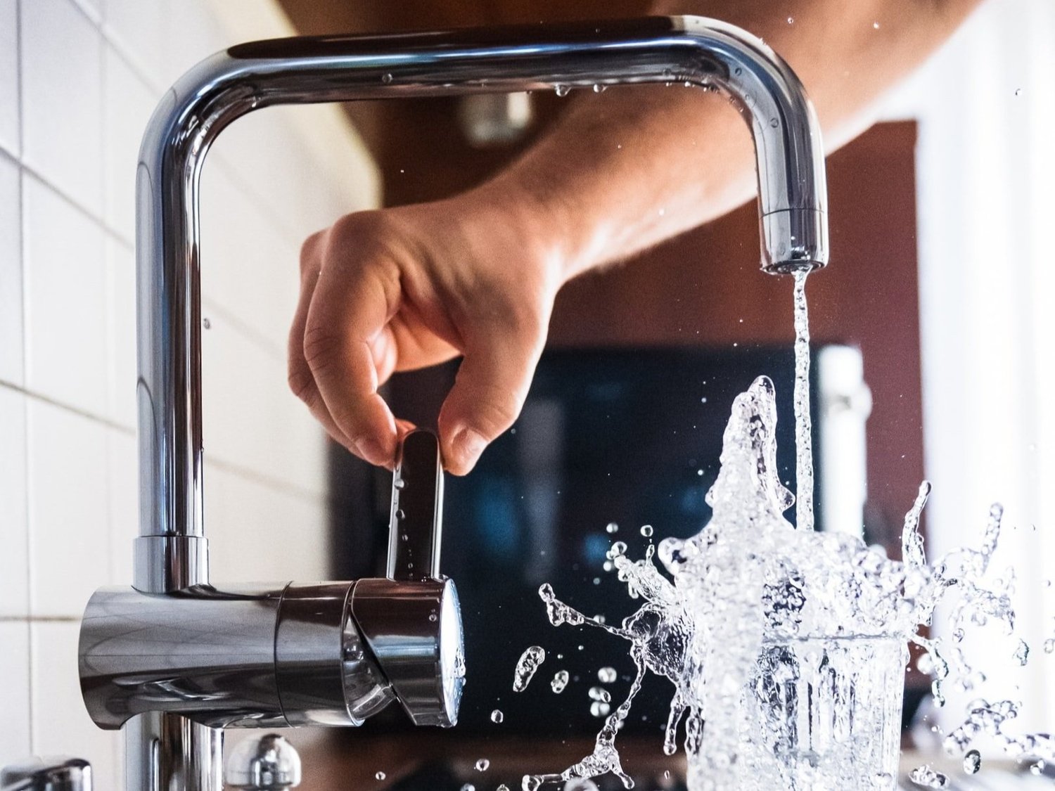 Hand turning on a modern chrome faucet with water splashing out of a glass beneath.