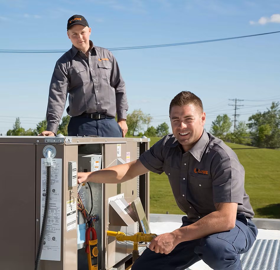 Two technicians wearing Lowe Mechanical Services uniforms fixing an air conditioning unit.