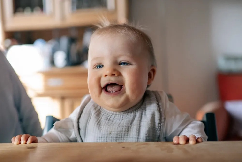 Ein lachendes Baby mit blauen Augen am Küchentisch.