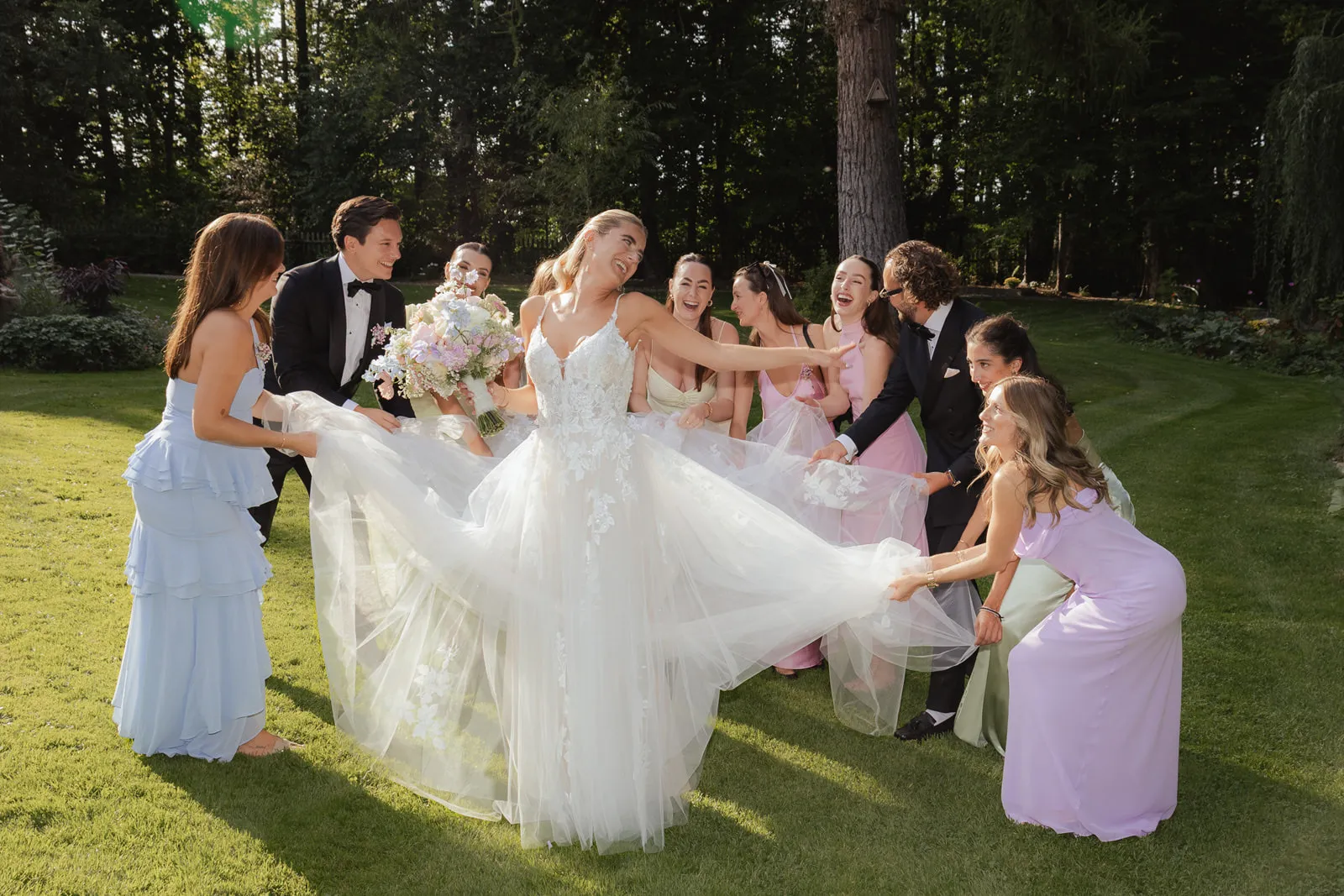 Braut in weißem Kleid lacht mit Bridesmaids und hält ihren Rock bei sonniger Gartenhochzeit auf hofgut Sickertshofen.