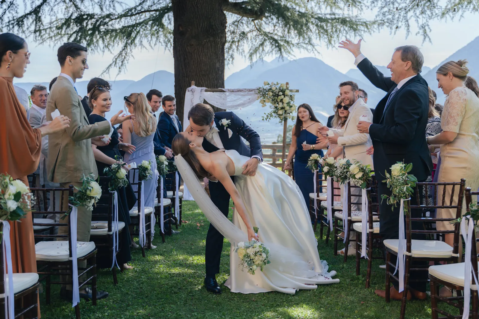 Brautpaar küsst sich nach Hochzeit im Freien in Meran, umgeben von applaudierenden Gästen und Blumen.