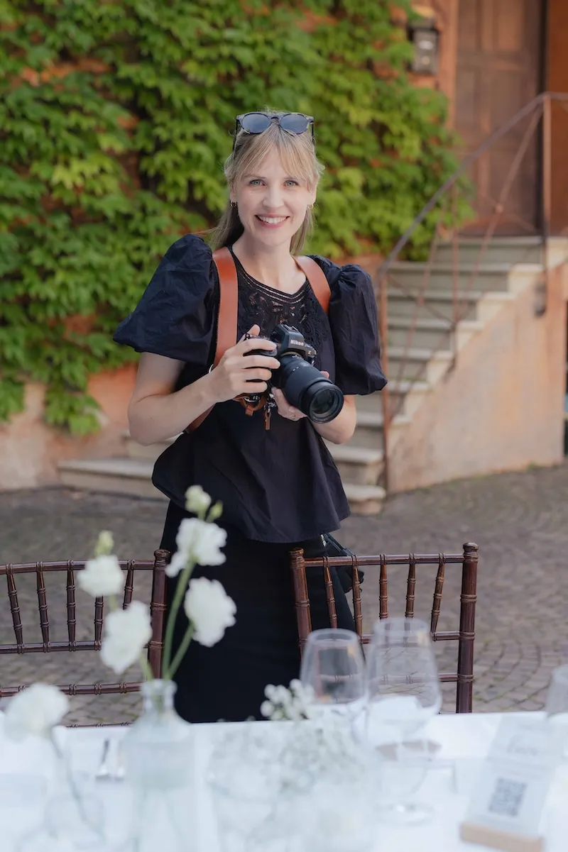 Stephanie Haberl mit Kamera, die lächelt, steht vor einer grünen Wand in Südtirol bei einer Hochzeit und spielt mit einer sonnigen Atmosphäre.