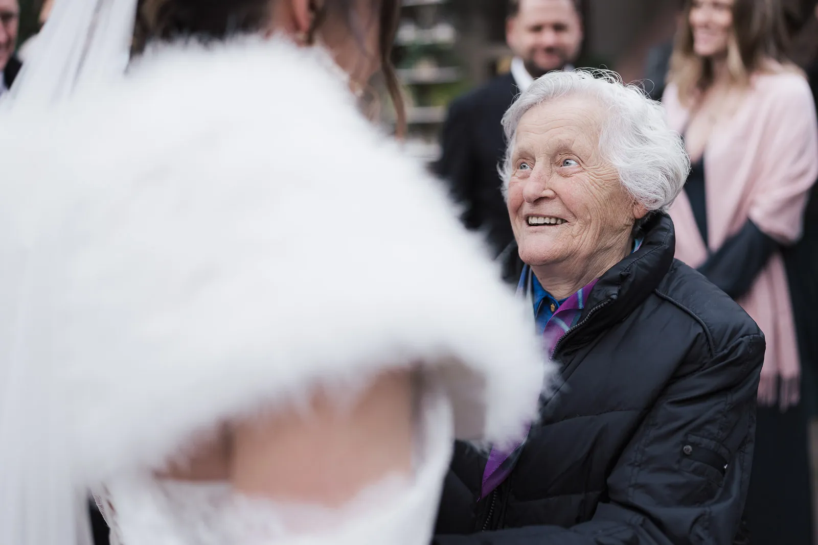 Ältere Frau mit weißen Haaren lächelt eine Person in einem weißen Kleid an, Hochzeit im Hintergrund.