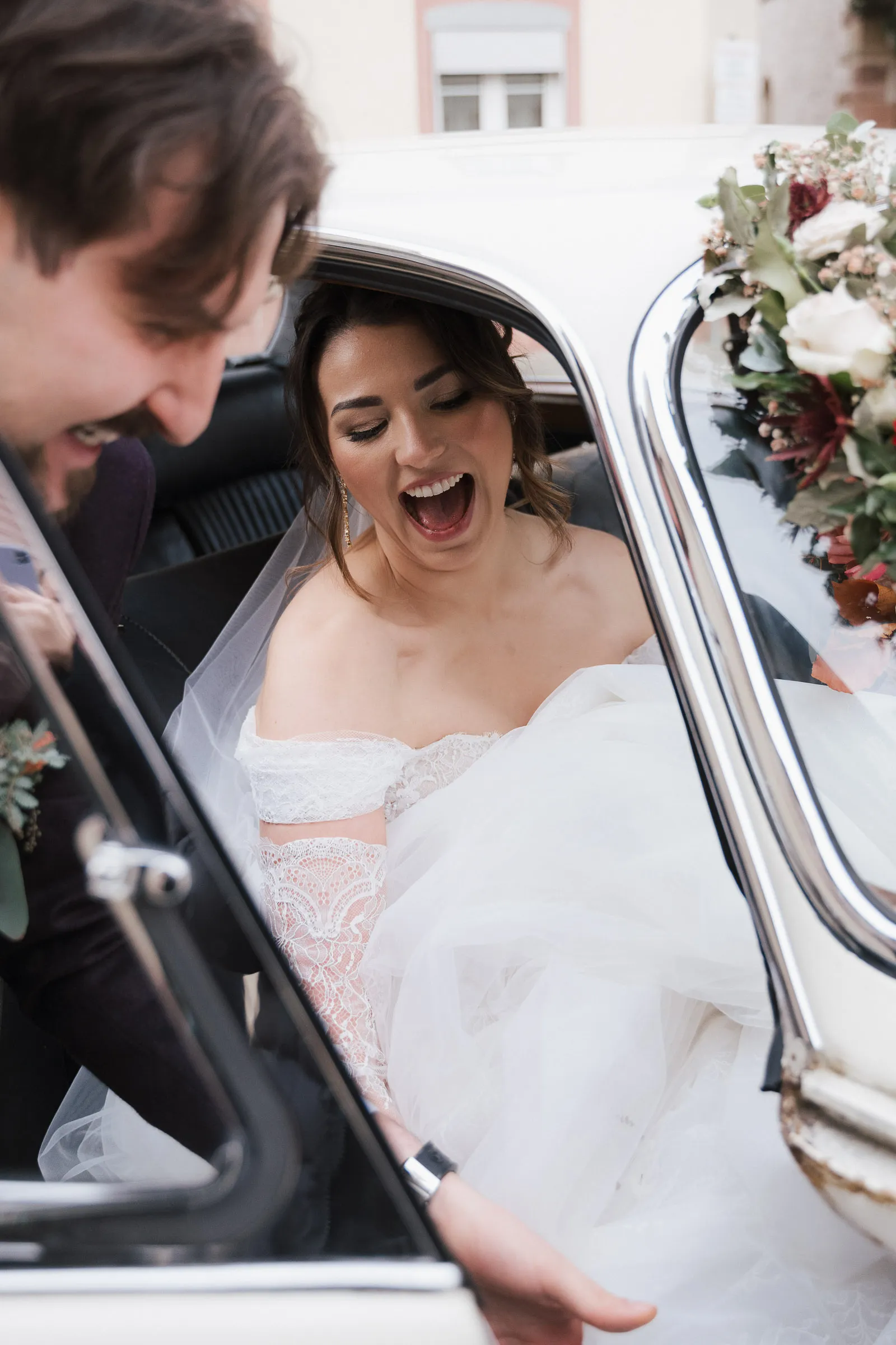 Glückliche Braut in Spitzenkleid steigt lachend in einen Oldtimer mit Blumenschmuck ein.