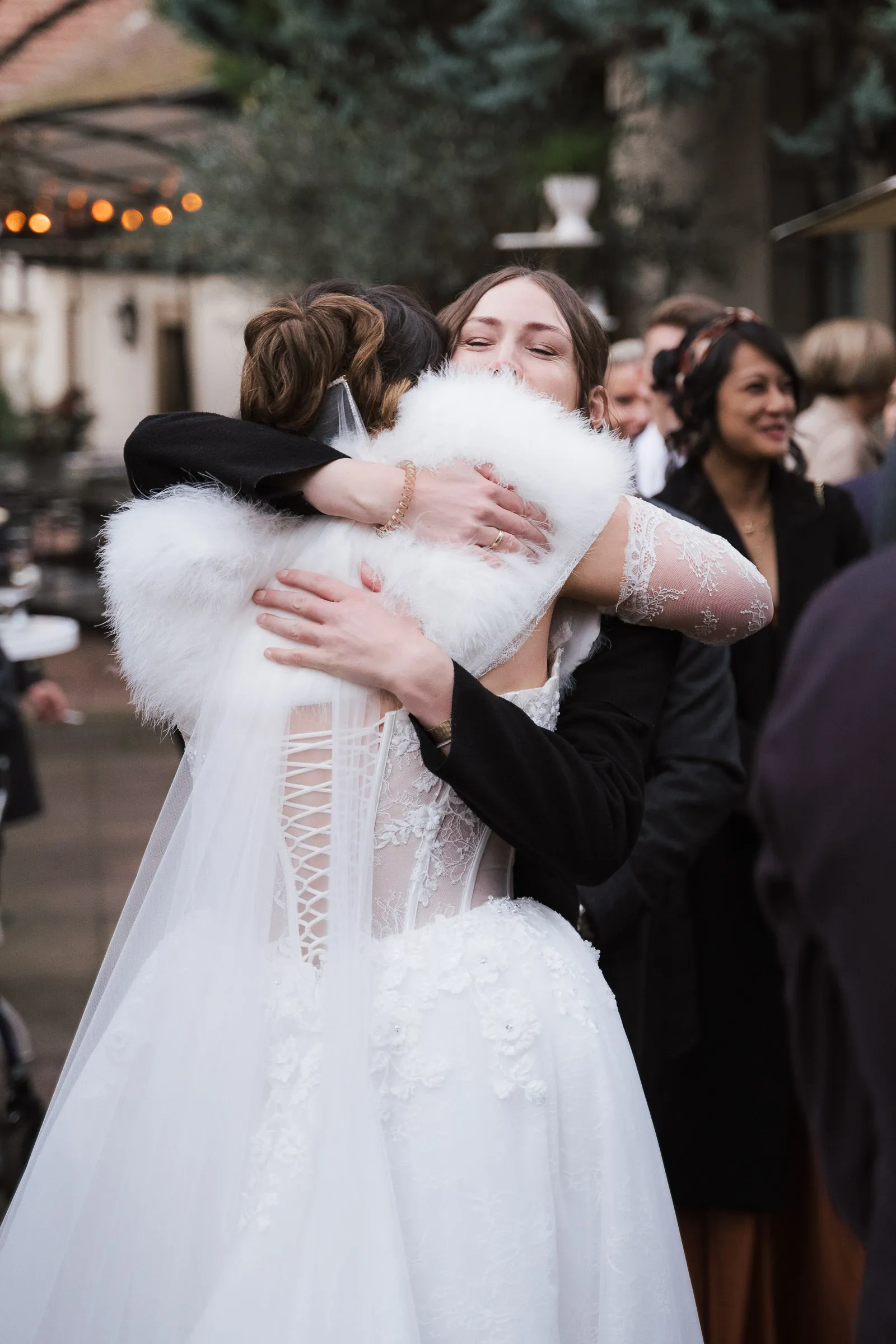 Braut in weißem Kleid mit Spitzenrücken umarmt lächelnde Frau bei Hochzeit mit Gästen im Hintergrund.