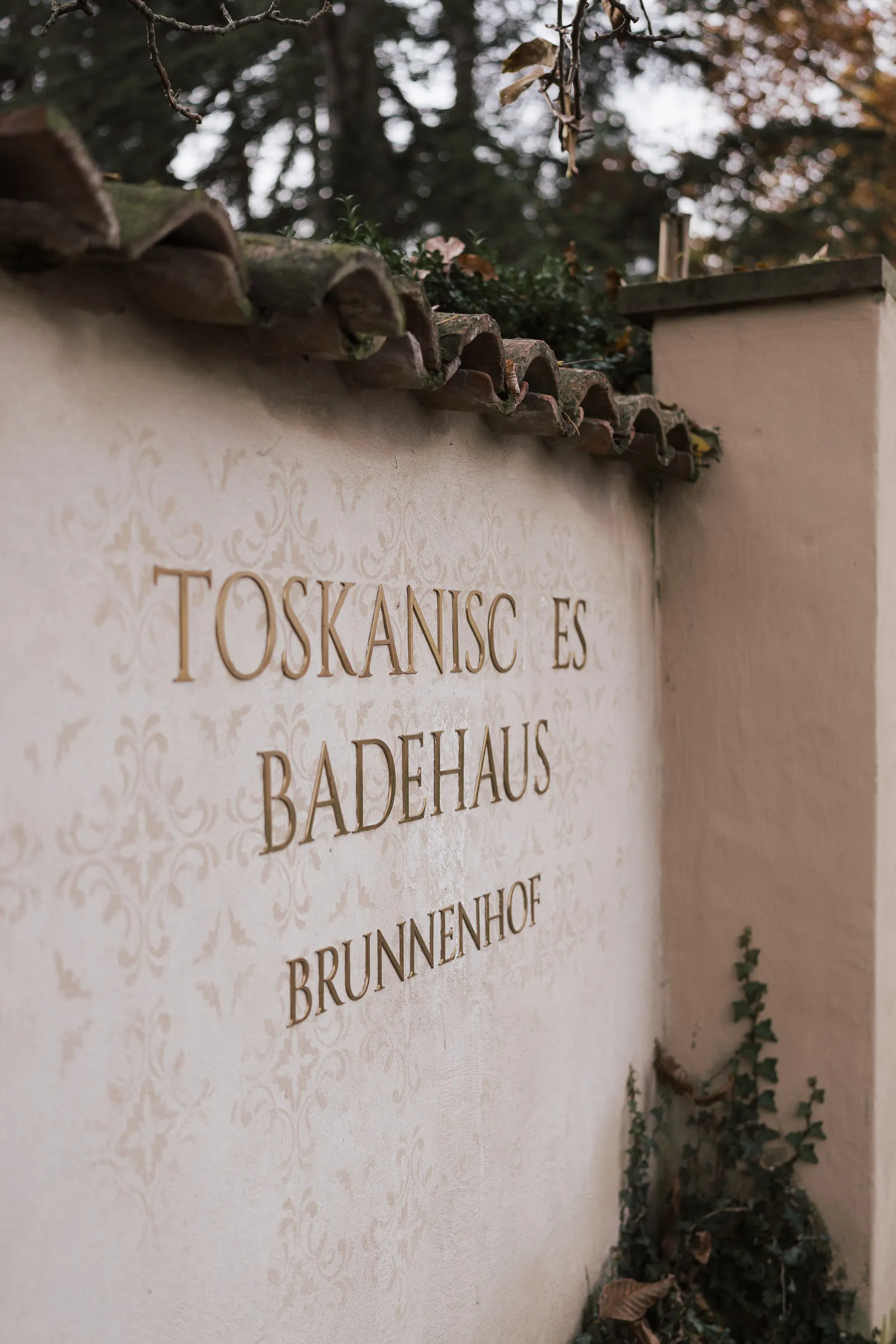 Beschriftete Mauer mit dem Schriftzug "Toskanisches Badehaus Brunnenhof" und Pflanzen am Rand.