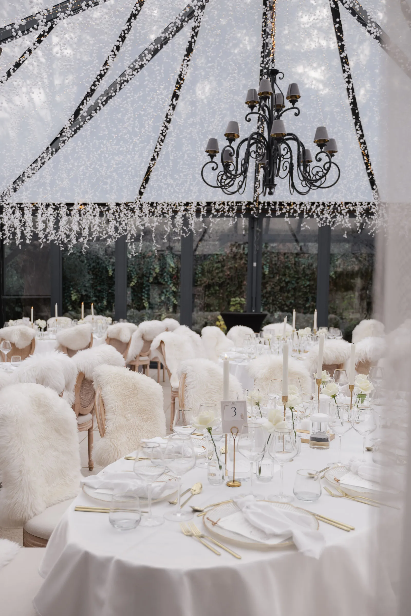 Elegante Winter-Hochzeitstische mit weißen Pelzbezügen, Kerzen und weißen Blumen in Zelt mit Kronleuchter in der Hochzeitslocation Morrhof.