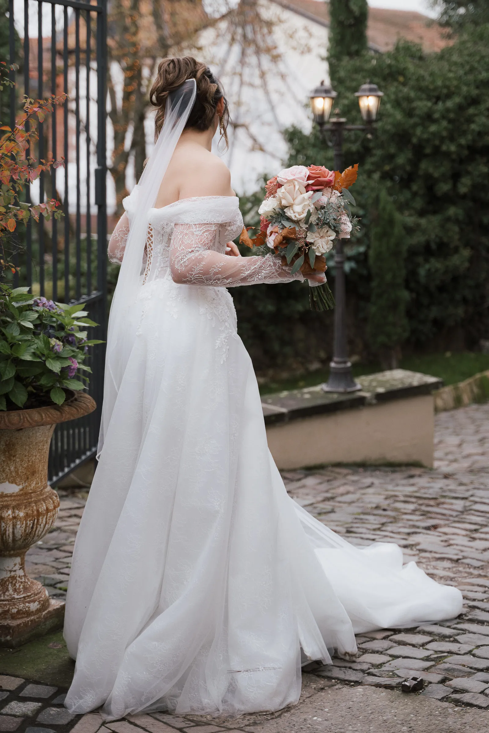 Braut in Spitzen-Hochzeitskleid mit Schleier hält Blumenstrauß, steht auf Kopfsteinpflaster im Morrhof.