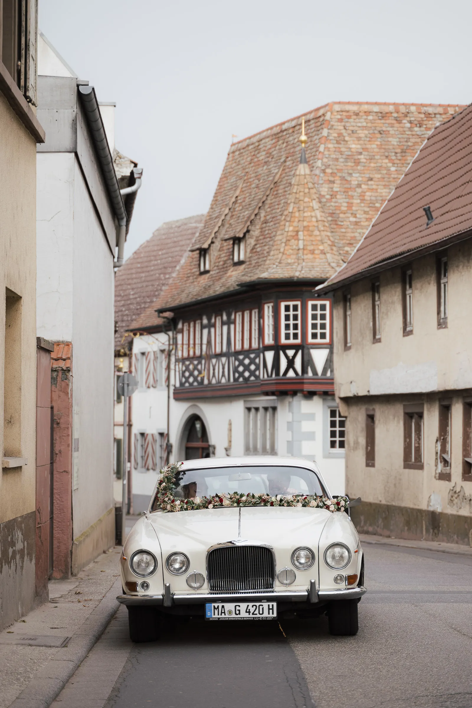 Weißer Oldtimer als Hochzeitsauto mit Blumenschmuck fährt durch enge Straße mit Fachwerkhäusern in Großkarlbach.