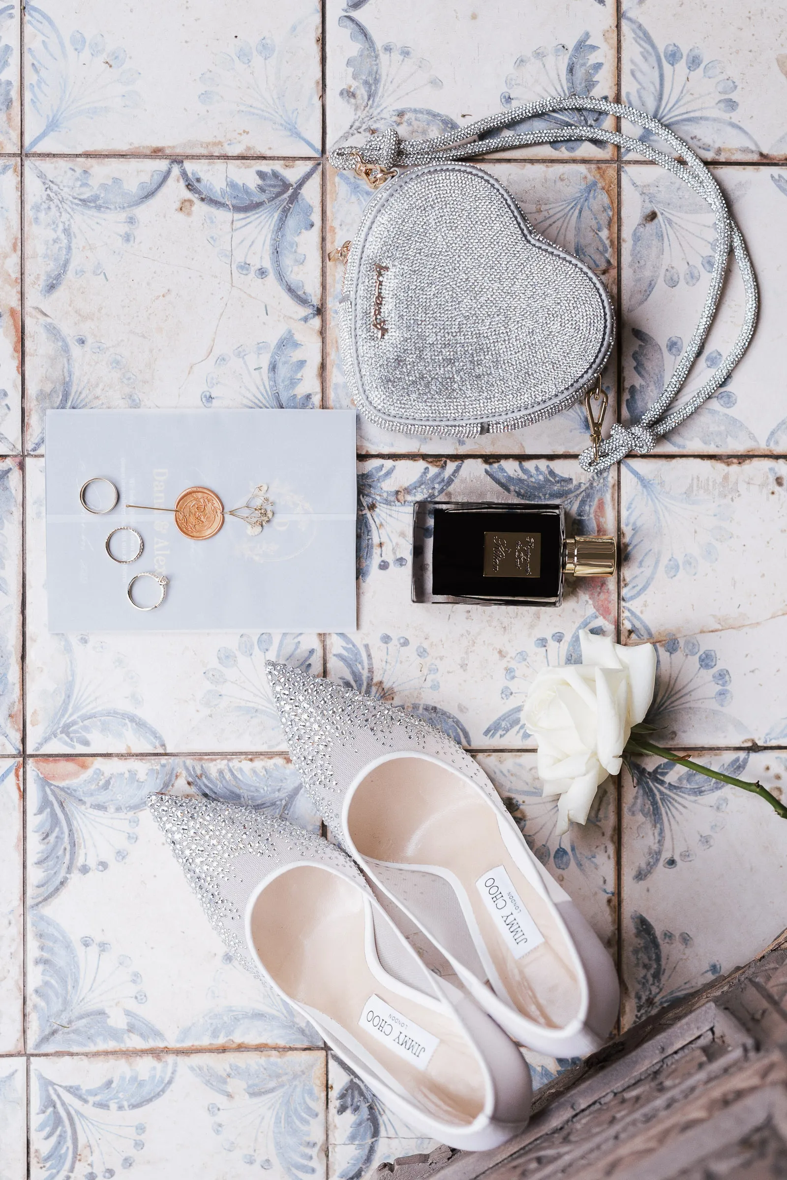 Brautschmuck, Jimmy Choo Schuhe, herzförmige Tasche, Parfum und weiße Rose auf gemusterten Fliesen.