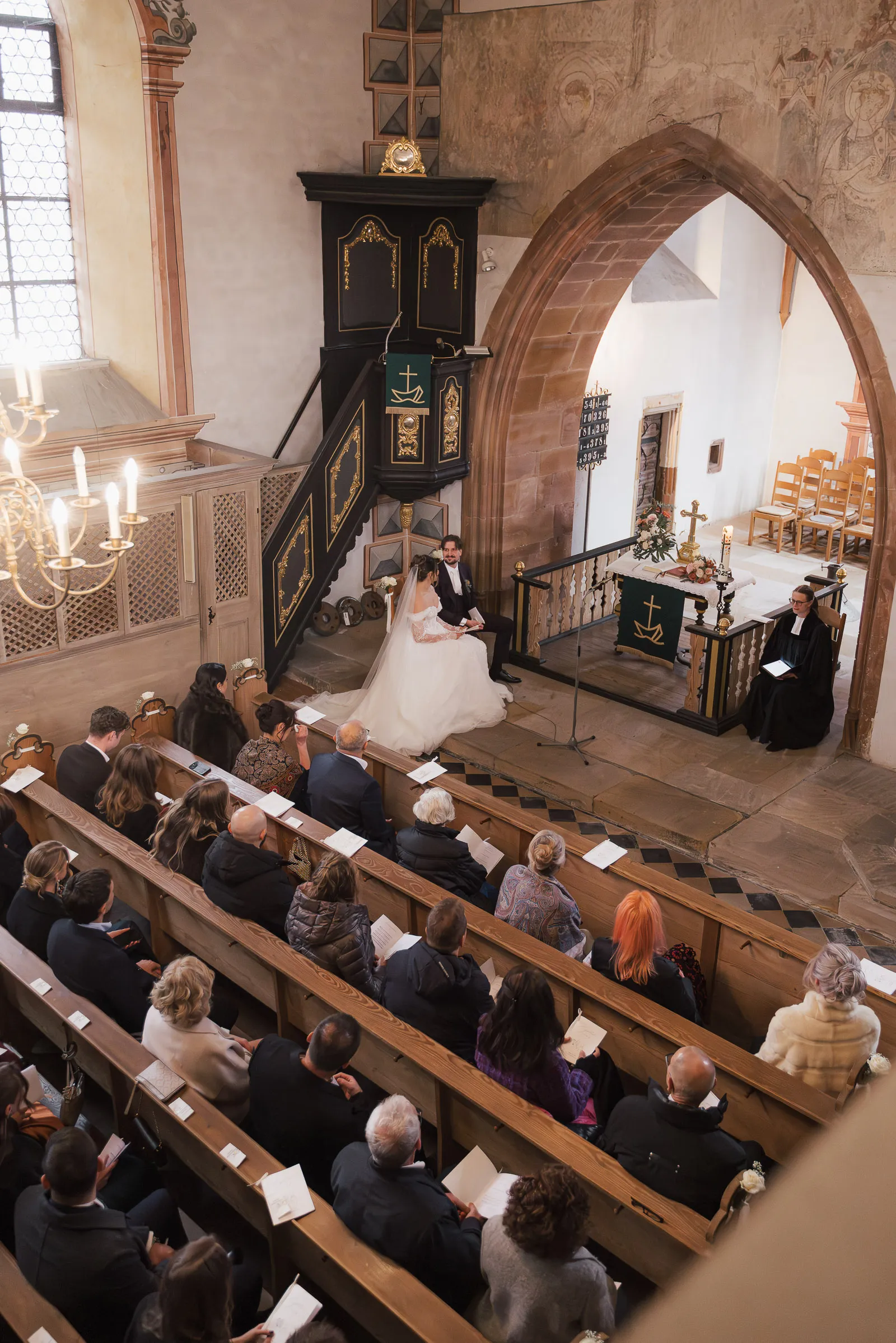 Brautpaar sitzt in der Kirche vor einem Geistlichen, Gäste sitzen auf Bänken und hören zu.